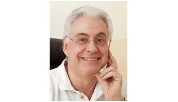 Dr. Herwig Bauer in weißem Polohemd und Brille lächelt freundlich, mit der Hand am Gesicht, vor einem hellen Hintergrund.