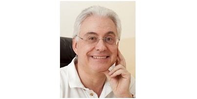 Dr. Herwig Bauer in weißem Polohemd und Brille lächelt freundlich, mit der Hand am Gesicht, vor einem hellen Hintergrund.