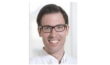 Dr. Philipp Becker in weißem Arztkittel und Brille lächelt vor einem hellen, unscharfen Hintergrund.
