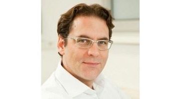 Dr. Albert Chavanne in weißem Hemd und Brille lächelt leicht vor einem hellen, modernen Praxis-Hintergrund.