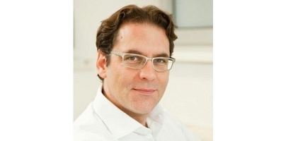 Dr. Albert Chavanne in weißem Hemd und Brille lächelt leicht vor einem hellen, modernen Praxis-Hintergrund.