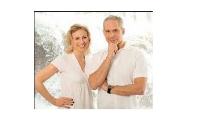 Ein Mann und eine Frau in weißer Kleidung stehen vor einem hellen Hintergrund. Die Frau lächelt, während der Mann nachdenklich seine Hand ans Kinn legt.