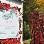 Praxis-Schild der Diagnosepraxis Dr. Schamp-Hertling, umrahmt von roten Herbstblättern, mit blauem Himmel und grünen Bäumen im Hintergrund.