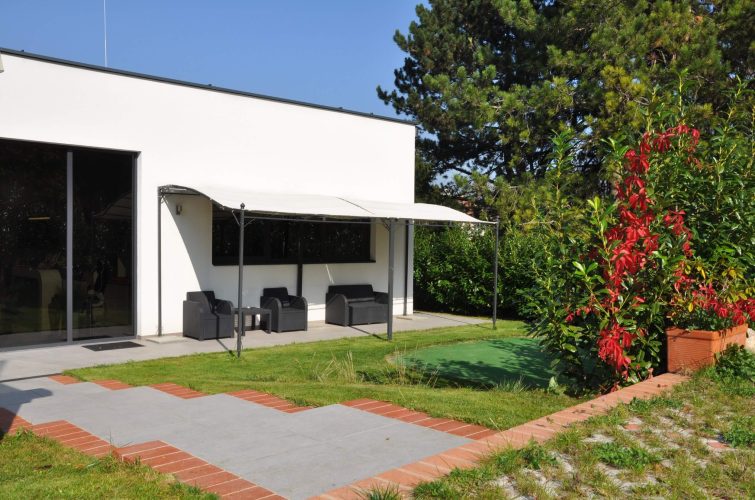 Der Außenbereich eines modernen Gebäudes, das als Diagnosepraxis dient. Ein Gehweg aus roten und grauen Platten führt zu einem Sitzbereich mit Gartenmöbeln unter einem Sonnenschutz. Im Hintergrund ist ein kleiner Golfplatz sichtbar, umgeben von grüner Vegetation und Bäumen. Der Himmel ist blau und wolkenlos.
