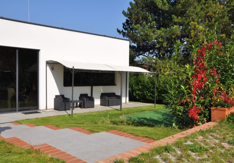 Der Außenbereich eines modernen Gebäudes, das als Diagnosepraxis dient. Ein Gehweg aus roten und grauen Platten führt zu einem Sitzbereich mit Gartenmöbeln unter einem Sonnenschutz. Im Hintergrund ist ein kleiner Golfplatz sichtbar, umgeben von grüner Vegetation und Bäumen. Der Himmel ist blau und wolkenlos.