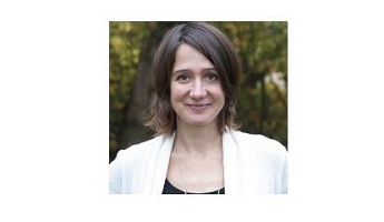 Dr. Ines Hertling in weißer Jacke und schwarzem Oberteil lächelt vor einem natürlichen, grünen Hintergrund.