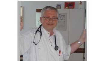 Dr. Rudolf Honetz, ein Arzt mit Brille und Stethoskop, steht lächelnd in einer Praxis. Im Hintergrund sind eine Sehtafel und medizinische Ausstattung zu sehen.