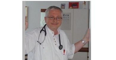 Dr. Rudolf Honetz, ein Arzt mit Brille und Stethoskop, steht lächelnd in einer Praxis. Im Hintergrund sind eine Sehtafel und medizinische Ausstattung zu sehen.