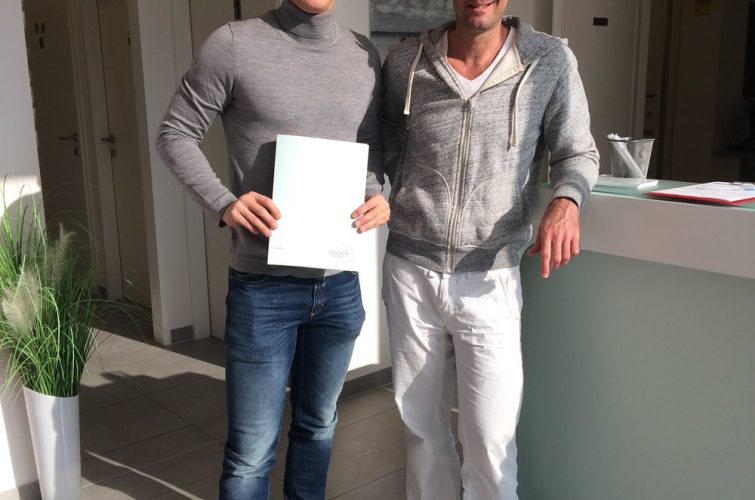 Dr. Med. Univ. Stephan Schamp-Hertling in grauer Kapuzenjacke und weißer Hose steht neben Roland Linz, der eine Mappe hält. Beide lächeln in einer modernen, hellen Praxis mit Rezeption und Pflanzen im Hintergrund.
