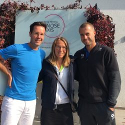 Dr. Med. Univ. Stephan Schamp-Hertling, Oliver Marach und eine Frau stehen lächelnd vor einem Praxisschild mit rotem Logo, umrahmt von Kletterpflanzen.