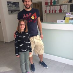 Jürgen Melzer in dunklem T-Shirt und beigen Shorts steht lächelnd neben einem Mädchen mit Brille und schwarzem Pullover in einer modernen Praxis mit Rezeption und Ordnerregalen im Hintergrund.