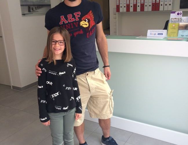 Jürgen Melzer in dunklem T-Shirt und beigen Shorts steht lächelnd neben einem Mädchen mit Brille und schwarzem Pullover in einer modernen Praxis mit Rezeption und Ordnerregalen im Hintergrund.