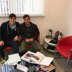 Sebastian Ofner und Gerald Melzer sitzen entspannt auf roten Sesseln im Wartebereich einer Praxis. Vor ihnen liegt ein Tisch mit Dokumenten, Magazinen und einem Spiel, im Hintergrund eine Kaffeemaschine und ein Fenster mit Jalousien.