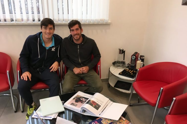 Sebastian Ofner und Gerald Melzer sitzen entspannt auf roten Sesseln im Wartebereich einer Praxis. Vor ihnen liegt ein Tisch mit Dokumenten, Magazinen und einem Spiel, im Hintergrund eine Kaffeemaschine und ein Fenster mit Jalousien.