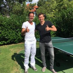 Dr. Med. Univ. Stephan Schamp-Hertling in weißer Kleidung steht neben Alexander Peya in schwarzem T-Shirt und grauer Jeans, beide zeigen den Daumen hoch. Im Hintergrund eine Tischtennisplatte, grüne Hecken und Bäume.
