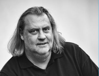 Schwarz-Weiß-Porträt von Dr. Richard Kdolsky mit schulterlangen Haaren und Bart, in schwarzem Polohemd, leicht lächelnd vor neutralem Hintergrund.