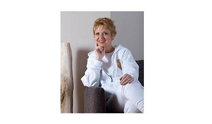 Dr. Andrea Schweitzer-Ehrenreich in weißer Kleidung sitzt entspannt auf einem Sessel, lächelnd mit der Hand am Kinn, vor einem schlichten Hintergrund.