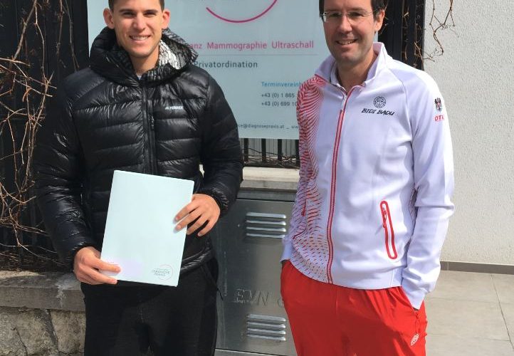 Dominic Thiem in schwarzer Jacke hält eine Mappe, während er neben Dr. Med. Univ. Stephan Schamp-Hertling in rot-weißer Sportkleidung vor einem Praxisschild steht.