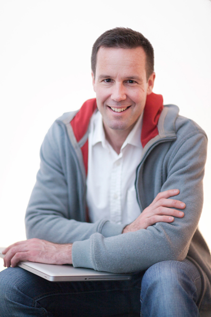Porträt von Dr. Stephan Schamp-Hertling mit Laptop Dr. Stephan Schamp-Hertling sitzt auf dem Boden, lächelt freundlich, trägt einen grauen Hoodie mit rotem Innenfutter und ein weißes Hemd. Er hält einen Laptop auf seinen Knien und schaut in die Kamera. Der Hintergrund ist hell und unscharf.