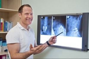 Dr. Andreas Kröner in weißem Polohemd steht lächelnd vor einem Leuchtkasten mit Röntgenbildern und zeigt mit einem Stift darauf, im Hintergrund ein Bücherregal.
