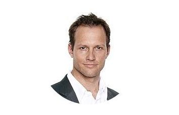 Dr. Lukas Brandner in dunklem Sakko und weißem Hemd blickt freundlich in die Kamera, vor einem weißen Hintergrund.