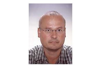 Dr. Alexander Kmen mit Brille und kurz rasiertem Haar trägt ein kariertes Hemd und blickt neutral in die Kamera vor einem schlichten Hintergrund.
