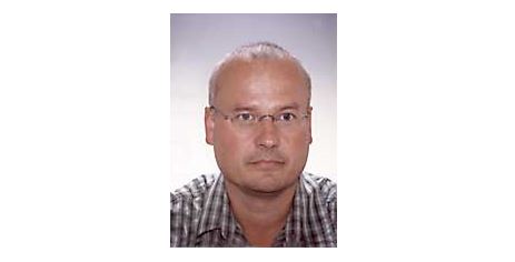 Dr. Alexander Kmen mit Brille und kurz rasiertem Haar trägt ein kariertes Hemd und blickt neutral in die Kamera vor einem schlichten Hintergrund.