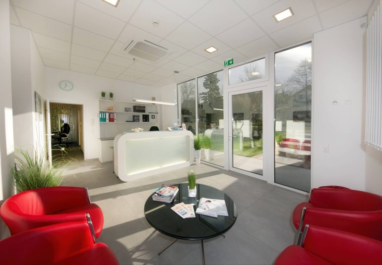 Heller und moderner Empfangsbereich der MRT-Diagnosepraxis mit roten Sesseln, Glastisch und Blick ins Freie.