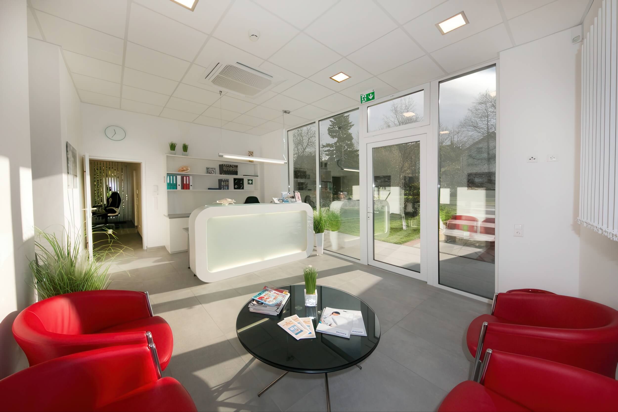 Heller und moderner Empfangsbereich der MRT-Diagnosepraxis mit roten Sesseln, Glastisch und Blick ins Freie.