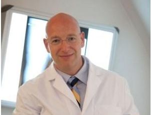 Dr. Christoph Müller in weißem Arztkittel und Krawatte lächelt in einer Praxis, im Hintergrund eine beleuchtete Röntgenaufnahme.