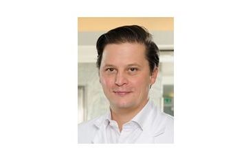 Dr. Paul Stampfl in weißem Arztkittel steht vor einem modernen, hellen Hintergrund und blickt freundlich in die Kamera.