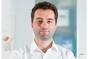 Dr. Aram Scherafati in weißem Hemd steht mit verschränkten Armen in einer modernen Praxis, im Hintergrund medizinische Poster und eine Glastür.