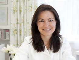Dr. Nicole Kotzailias in weißem Arztkittel sitzt lächelnd in einer hellen Praxis mit Blumen und Vorhängen im Hintergrund.