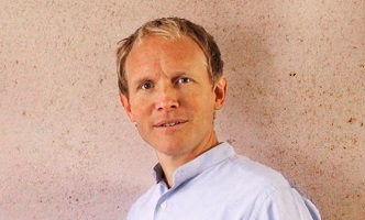 Dr. Thomas Wögerbauer in hellblauem Hemd steht vor einer beige strukturierten Wand und blickt freundlich in die Kamera.