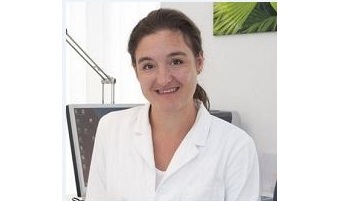Dr. Caroline Pohl in weißem Arztkittel lächelt in einer modernen Praxis, im Hintergrund ein Schreibtisch mit Computer und eine grüne Wanddekoration.