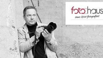 Schwarz-Weiß-Bild von Fotograf Hans Krist, der mit einer Kamera an einer Wand lehnt. Oben rechts das Logo von „foto haus“ mit seinem Namen.