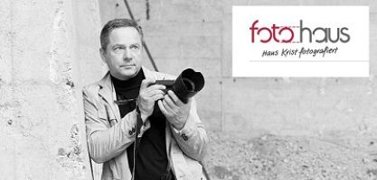 Schwarz-Weiß-Bild von Fotograf Hans Krist, der mit einer Kamera an einer Wand lehnt. Oben rechts das Logo von „foto haus“ mit seinem Namen.