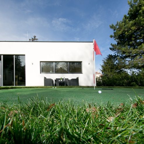 Eine Aufnahme eines Golfplatzes mit einem grünen Rasen im Vordergrund und einem roten Fahnenmast. Im Hintergrund ist ein modernes Gebäude zu sehen, das als Diagnosepraxis dient. Durch die Fenster des Gebäudes sind Möbel erkennbar. Der Himmel ist klar, und es gibt einen Baum auf der rechten Seite des Bildes.