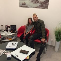 Marko Arnautović in dunkler Kleidung sitzt neben einem lächelnden Mädchen auf roten Sesseln in einer Praxis. Im Vordergrund ein Glastisch mit Dokumenten, im Hintergrund eine Kaffee-Ecke und ein Landschaftsbild an der Wand.